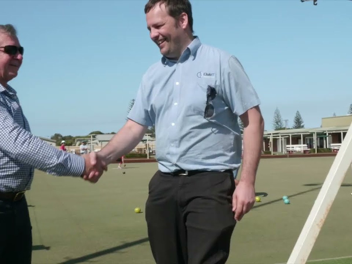 A ClubIT engineer in a branded shirt shaking hands
               with the chief executive of Evans Head Bowling Club on the club's bowling green, with the
               clubhouse and players behind them